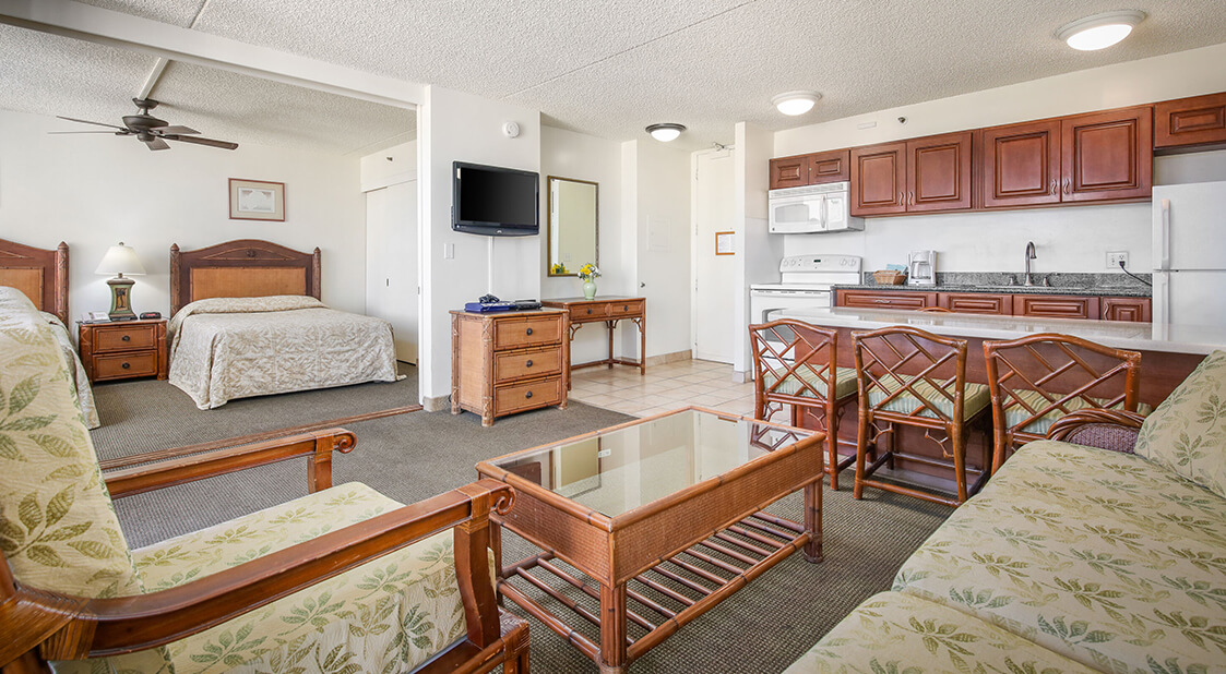 Living room with a couch, coffee table, TV, and entry to kitchen and bedroom with 2 beds.