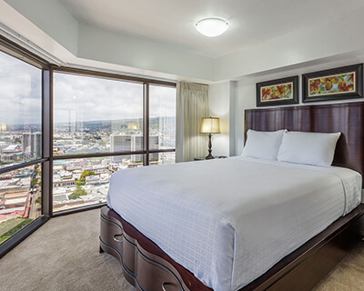 Room with a bed, lamp, and large picture windows with city and ocean views.