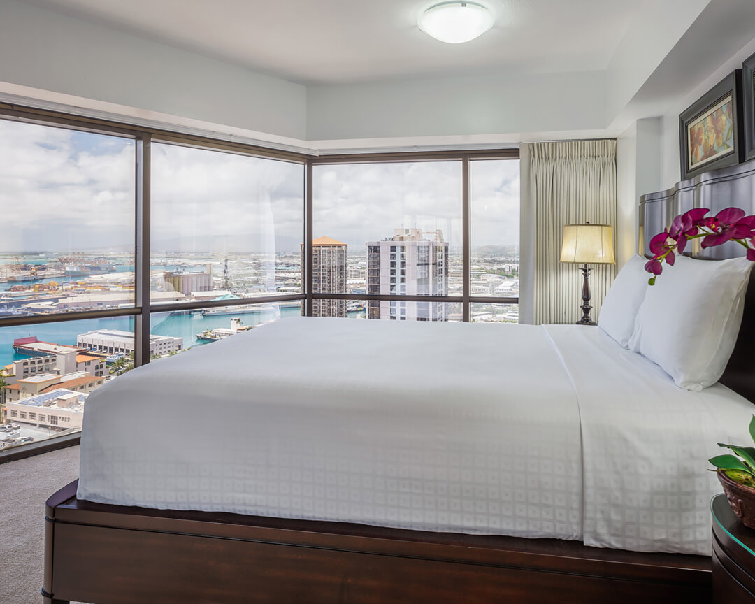 Room with a bed, lamp, and large picture windows with city and ocean views.