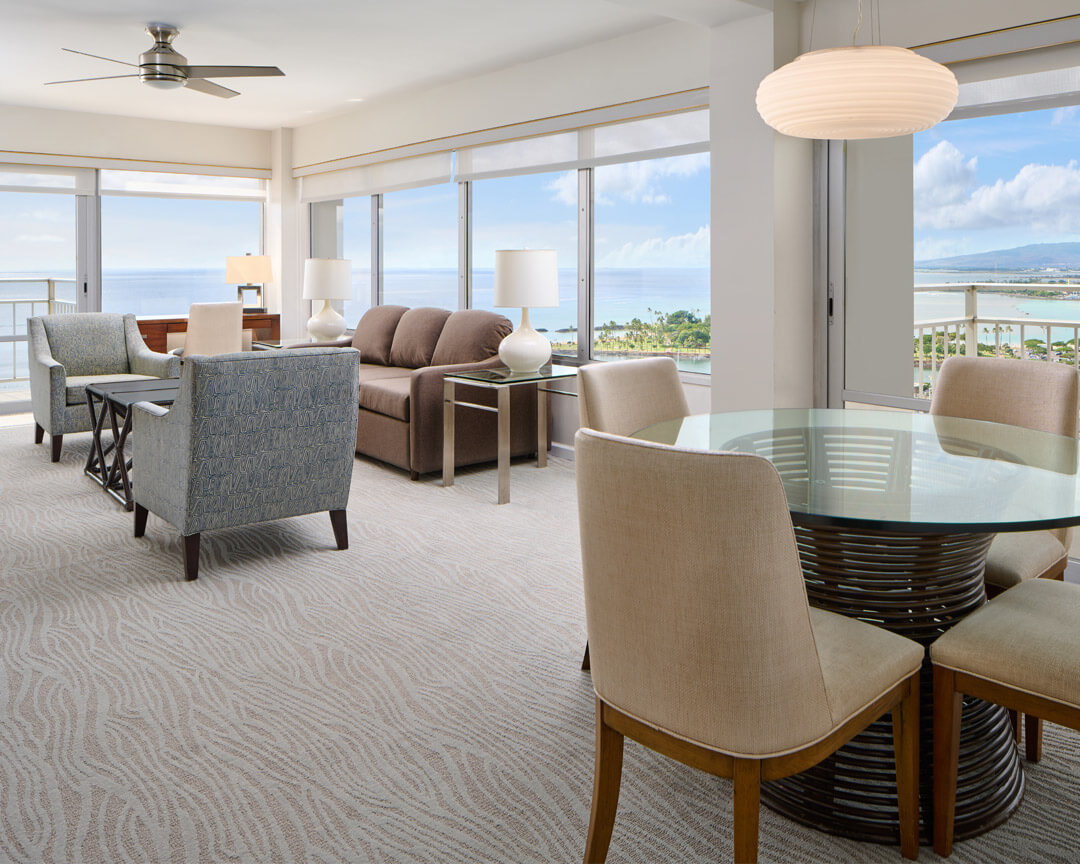Living and dining area with floor to ceiling windows along two walls, overlooking the ocean.