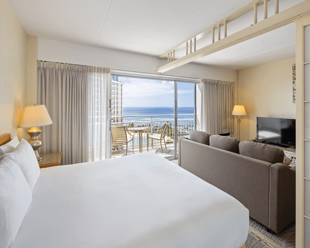 Bedroom with sofa, large windows and balcony overlooking the ocean.