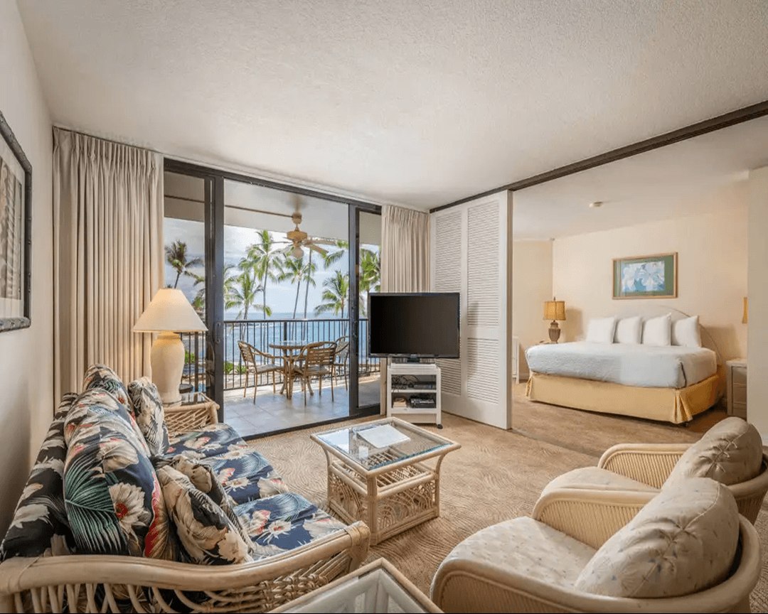 Living area with couch, coffee table, TV, balcony overlooking the ocean.