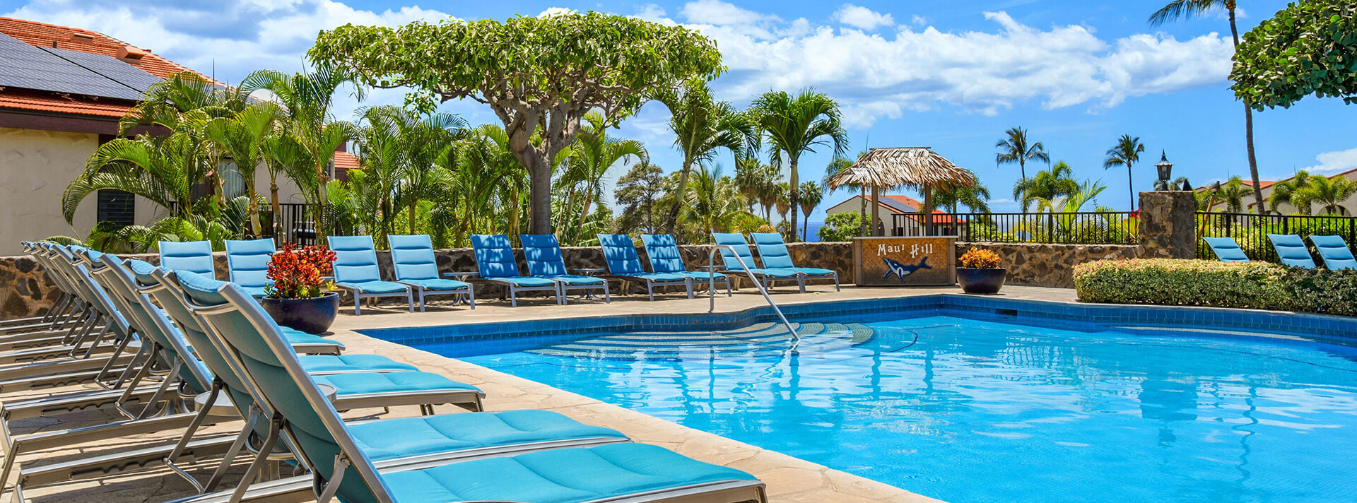 Pool at the Maui Hill with blue padded lounge chairs, landscaped with lush green palm trees and colorful bushes and accommodation building units in the background