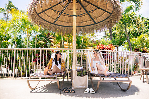 2 women relaxing on loungers under grass hut shade